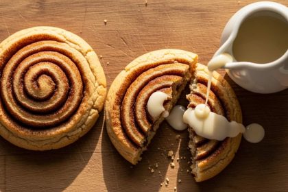 Overhead shot of soft, swirled cinnamon roll cookies with a broken piece and a drizzle of cream cheese glaze.